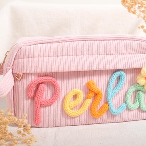 Peut inclure: Une trousse de maquillage en velours côtelé rose avec le nom "Perla" en lettres tricotées colorées. Le sac a une poignée et une fermeture éclair. Une fleur tricotée est à côté du nom. Le sac est sur une surface blanche.