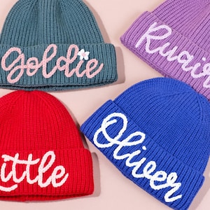 May include: Four colourful knitted beanies are displayed. The beanies are teal, red, purple, and blue. Each beanie has a name embroidered in a contrasting colour: "Goldie", "Little", "Ruairi", and "Oliver". The beanies are a cosy and stylish accessory.