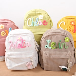 Peut inclure: Cinq sacs à dos en velours côtelé rose, blanc, jaune, vert et marron. Chaque sac à dos porte un nom brodé en fil coloré, comme "Perl" et "Chloe", et une fleur décorative. Les sacs à dos ont une poche avant et des bretelles réglables.
