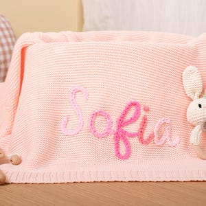 May include: A light pink knitted baby blanket with the name "Sofia" embroidered in pink. A crochet bunny rattle and a wooden toy are on the blanket. The blanket is folded and placed on a wooden surface.