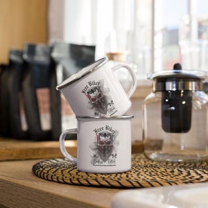 May include: Two white enamel mugs stacked on a woven mat. Each mug features a black and red graphic of a skull wearing a bandana, with the words "Free Biker" and "Badass Edition". The mugs have silver rims and white handles.