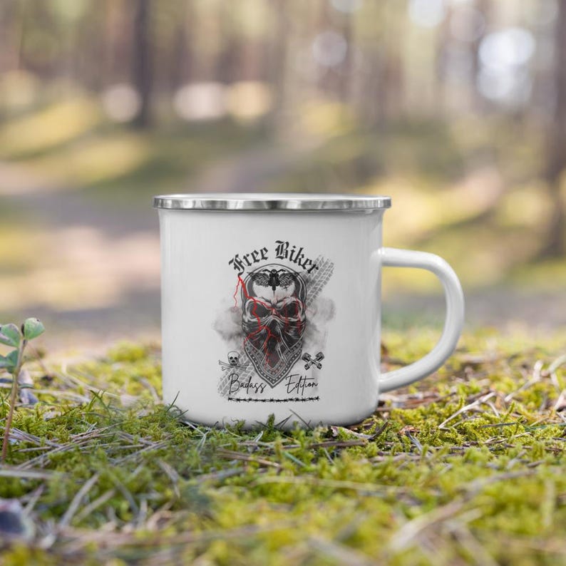 May include: White enamel mug with a silver rim featuring a skull design wearing a red bandana. The text "Free Biker Badass Edition" is printed on the mug. The mug has a white handle and is set against a blurred green background.