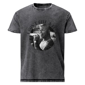 Könnte beinhalten: Ein graues T-Shirt mit Säurewaschung und einem schwarz-weißen kreisförmigen Grafik eines rauchenden Mannes. Die Grafik ist von dem Text "I DON'T GIVE A FUCK" umgeben. Das Shirt hat einen Rundhalsausschnitt und kurze Ärmel.