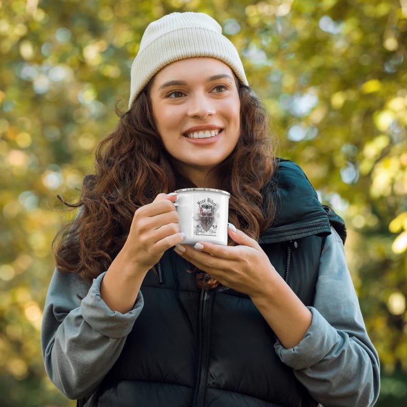 May include: White enamel mug with a black rim and a graphic design that includes the text "Beer Biker". The mug is held by a person wearing a white beanie and a black vest. The background is blurred with green and yellow tones.