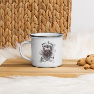 May include: White enamel mug with a black rim, featuring the text "Free Biker" and a skull design with a red bandana. The mug is on a wooden surface with cookies.