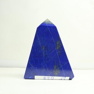 Può includere: Un obelisco di lapislazzuli blu a forma triangolare. La pietra presenta venature bianche e dorate visibili e una consistenza leggermente ruvida. La parte superiore dell'obelisco è di colore grigio chiaro. Una piccola etichetta bianca con scritta blu è attaccata alla base.