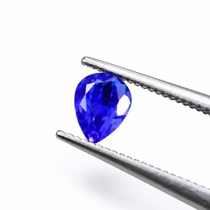 Op de afbeelding: Een peervormige, gefacetteerde blauwe edelsteen vastgehouden door een zilverkleurige metalen pincet. De steen heeft een levendige, diepblauwe kleur met meerdere interne facetten die het licht reflecteren, wat zorgt voor een briljante, sprankelende uitstraling.