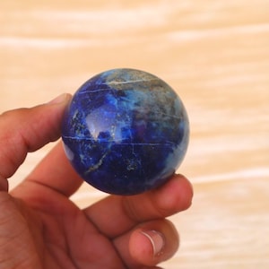 Op de afbeelding: Een gepolijste lapis lazuli bol, met een diepblauwe tint met lichtere blauwe en gouden aderen. De edelsteen wordt in een hand gehouden, tegen een lichtbruine achtergrond. De bol heeft een diameter van ongeveer 5 cm.