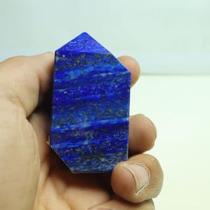 Può includere: Un obelisco di lapislazzuli blu, tenuto in mano. La pietra ha un colore blu intenso con striature di blu più chiaro e macchie dorate. L'obelisco è tagliato con una parte superiore e lati piatti.