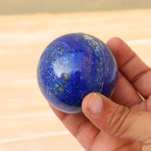 Puede incluir: Una esfera de lapislázuli azul profundo con motas doradas y blancas. La gema pulida se sostiene en una mano, mostrando su forma lisa y redondeada. El color de la esfera varía de un azul oscuro intenso a tonos más claros, con inclusiones minerales visibles.