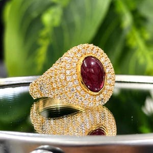 Puede incluir: Un anillo de oro con una piedra preciosa central de color rojo intenso en forma de cabujón ovalado. La superficie del anillo está densamente incrustada con numerosas piedras pequeñas, claras y de corte redondo que rodean la piedra central.