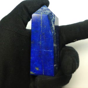 Può includere: Un obelisco di lapislazzuli blu intenso. La pietra lucidata presenta macchie e venature dorate naturali. L'obelisco è tenuto in una mano guantata di nero. La gemma ha una forma rettangolare con una parte superiore appuntita.