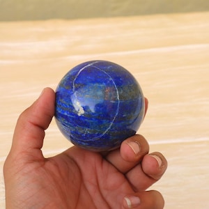 Può includere: Una sfera di lapislazzuli lucidata, tenuta in mano. La sfera è di un blu intenso con striature di blu più chiaro e oro. La pietra è liscia e rotonda, con una leggera lucentezza.