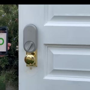 May include: A smart lock system with a gray exterior and a gold-colored doorknob is mounted on a white door. A smartphone displays a green circle, indicating the lock's status. The background includes green foliage.