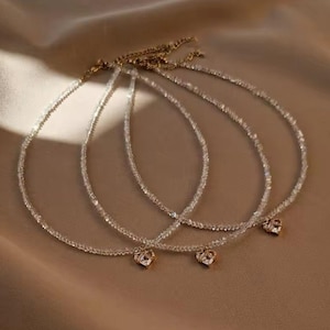 May include: Three delicate necklaces with clear, sparkling beads and small gold-colored heart pendants. The necklaces are arranged on a soft, neutral-toned fabric, creating a simple and elegant display. The necklaces are likely for women.