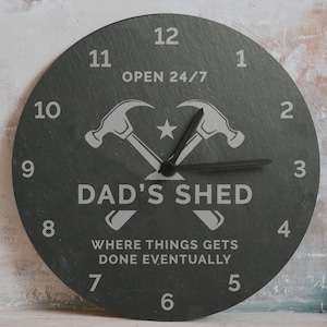 May include: A circular, dark grey clock with white numerals and hands. The clock face displays the text "OPEN 24/7", "DAD'S SHED", and "WHERE THINGS GETS DONE EVENTUALLY". Two crossed hammers and a star are also present.