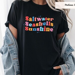 May include: A black short-sleeved t-shirt featuring the text Saltwater Seashells Sunshine in colorful, wavy, retro-style lettering. The design includes small blue star accents. A light blue denim jacket is draped over the shoulders of the shirt.