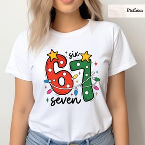 May include: White t-shirt with a colorful graphic design featuring the numbers "6" and "7" in red and green, adorned with star accents and string lights. The word "seven" is written below the numbers. The brand name "Meliona Veste" is at the top.