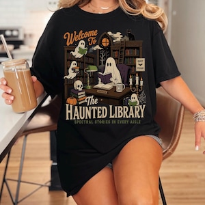 May include: A black t-shirt featuring a graphic of a library with ghosts, books, a skull, a pumpkin, and a cat. The text reads: Welcome To The HAUNTED LIBRARY SPECTRAL STORIES IN EVERY AISLE. A small sign on the desk says LIBRARIAN and a mug says READ IF YOU DARE.