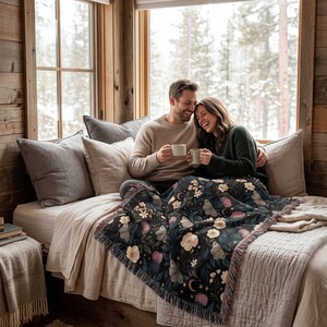 May include: A woven throw blanket featuring a dark background with a repeating pattern of light-colored flowers, moths, and crescent moons, finished with fringed edges, draped over a bed with neutral-toned pillows and bedding.