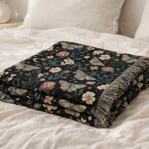May include: A folded woven throw blanket featuring a dark black background with a repeating pattern of light green moths, pink and white flowers, green foliage, small yellow stars, and a crescent moon, finished with a fringed edge on the side.