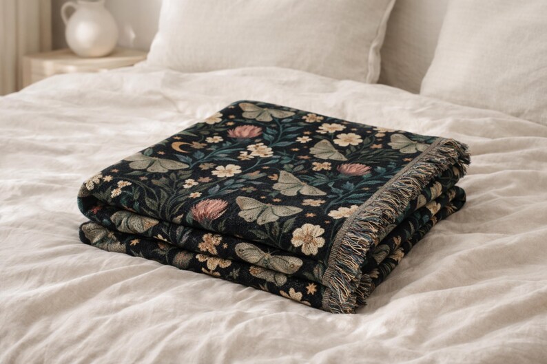May include: A folded woven throw blanket featuring a dark black background with a repeating pattern of light green moths, pink and white flowers, green foliage, small yellow stars, and a crescent moon, finished with a fringed edge on the side.
