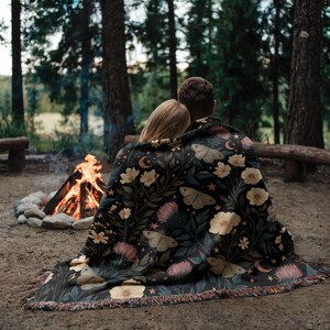 May include: A black woven throw blanket featuring a botanical pattern of cream-colored flowers, pink thistle-like blooms, and moths. The design includes small crescent moons and stars scattered throughout, finished with a fringed edge.
