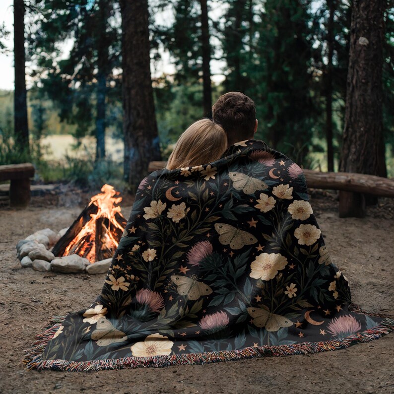 May include: A black woven throw blanket featuring a botanical pattern of cream-colored flowers, pink thistle-like blooms, and moths. The design includes small crescent moons and stars scattered throughout, finished with a fringed edge.