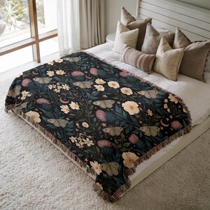 May include: A woven throw blanket featuring a dark background with a repeating pattern of moths, light-colored flowers, thistle-like plants, small crescent moons, and stars. The blanket has a fringed edge and is draped over a bed with several pillows.