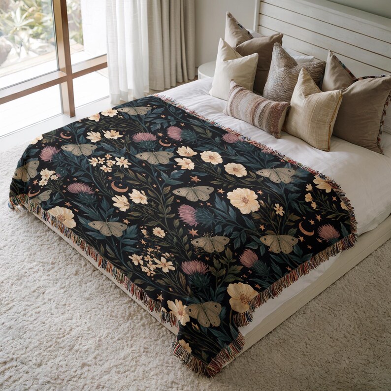 May include: A woven throw blanket featuring a dark background with a repeating pattern of moths, light-colored flowers, thistle-like plants, small crescent moons, and stars. The blanket has a fringed edge and is draped over a bed with several pillows.