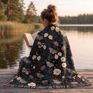 May include: A woven throw blanket featuring a dark background with a botanical pattern of white flowers, pink thistle-like blooms, moths, and crescent moons. The blanket has a fringed edge and is draped over the back of a person reading a book.