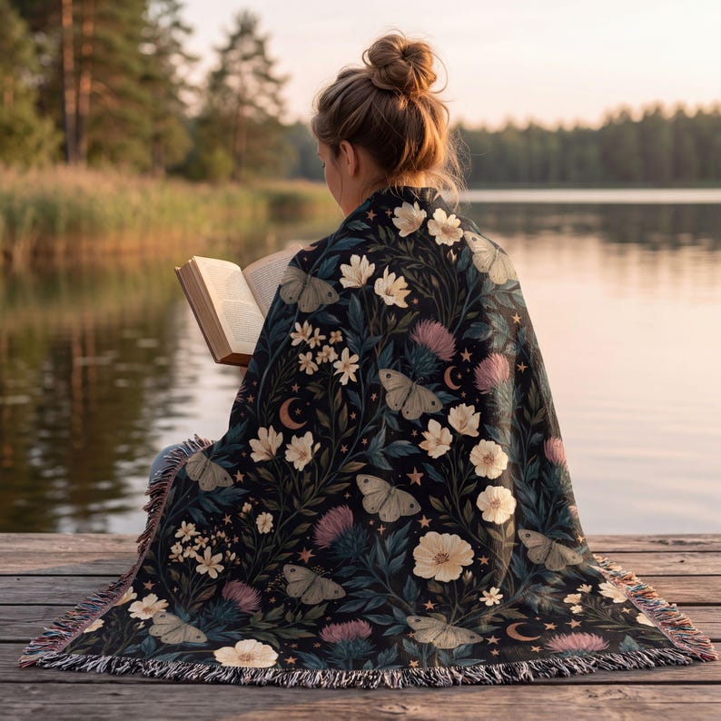 May include: A woven throw blanket featuring a dark background with a botanical pattern of white flowers, pink thistle-like blooms, moths, and crescent moons. The blanket has a fringed edge and is draped over the back of a person reading a book.