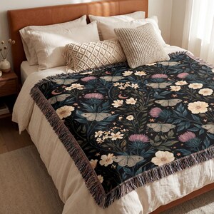 May include: A woven throw blanket featuring a dark black background with a repeating pattern of light-colored moths, cream-colored flowers, purple thistle-like blooms, small crescent moons, and tiny stars, finished with a decorative fringe border.