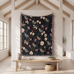 May include: A woven throw blanket featuring a dark black background with a repeating pattern of light-colored moths, cream-colored flowers, pink thistle-like blooms, and small crescent moons and stars. The blanket has a fringed edge on all sides.