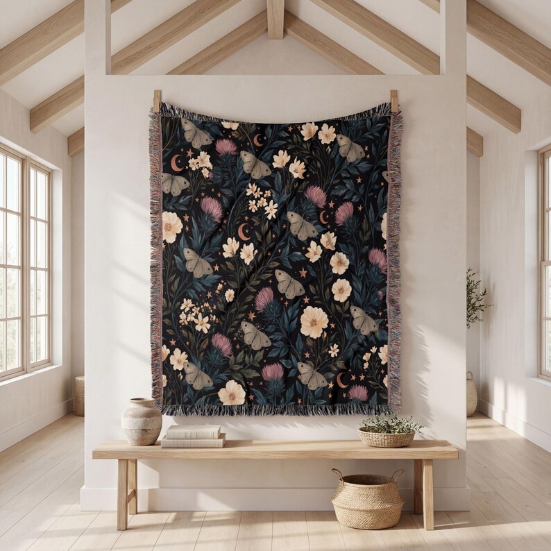 May include: A woven throw blanket featuring a dark black background with a repeating pattern of light-colored moths, cream-colored flowers, pink thistle-like blooms, and small crescent moons and stars. The blanket has a fringed edge on all sides.