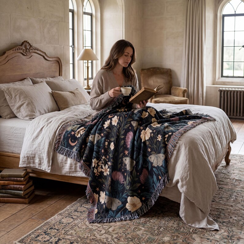 May include: A dark-colored woven throw blanket featuring a repeating pattern of cream-colored flowers, light-colored moths, crescent moons, and stars. The blanket has a fringed edge and is draped over a bed with light-colored bedding and pillows.