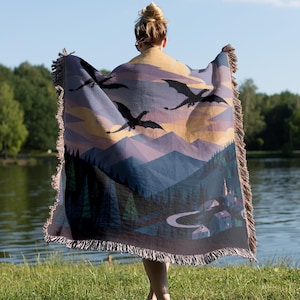 May include: A woven throw blanket featuring a landscape illustration of dark silhouettes of dragons flying over purple mountains, a dense forest of pine trees, and a small village with a winding road, finished with a decorative fringed edge.