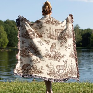 May include: A cream-colored woven throw blanket featuring a repeating brown pattern of woodland animals including deer, an owl, a fox, and hedgehogs, interspersed with mushrooms, oak leaves, and acorns, finished with a fringed edge on all sides.