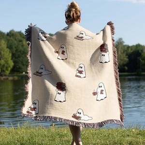 May include: A cream-coloured woven throw blanket featuring a repeating pattern of white ghosts. Some ghosts are reading books, some have stacks of books on their heads, and others are holding mugs. The blanket has fringed edges on all sides.