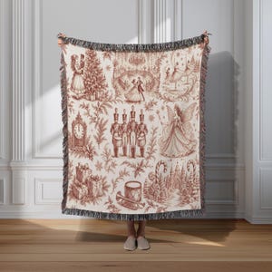 May include: A woven throw blanket featuring a cream background with a repeating deep red toile-style print depicting Nutcracker-themed scenes, including toy soldiers, a fairy, a clock, wrapped gifts, and dancers, finished with a fringed edge.
