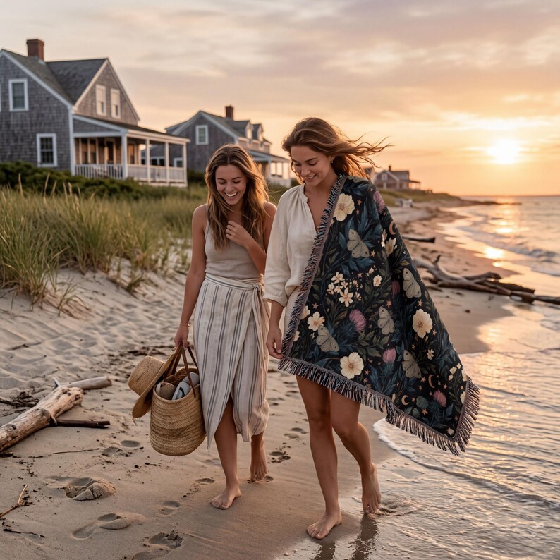 May include: A woven throw blanket featuring a dark navy background with a repeating pattern of cream-colored flowers, moths, and crescent moons, finished with a fringed edge. The blanket is draped over an arm while walking along a sandy beach.