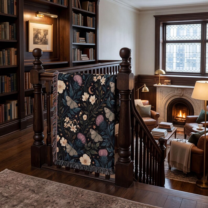 May include: A woven throw blanket featuring a dark background with a botanical pattern of white flowers, purple thistle, and light-colored moths. The design includes small stars and a crescent moon, finished with a fringed edge on the sides.