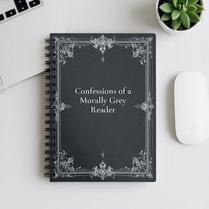 May include: A spiral-bound notebook with a dark gray cover featuring the text "Confessions of a Morally Grey Reader" in a decorative font. The cover has an ornate silver frame design. A white computer mouse and a small potted succulent are also visible.