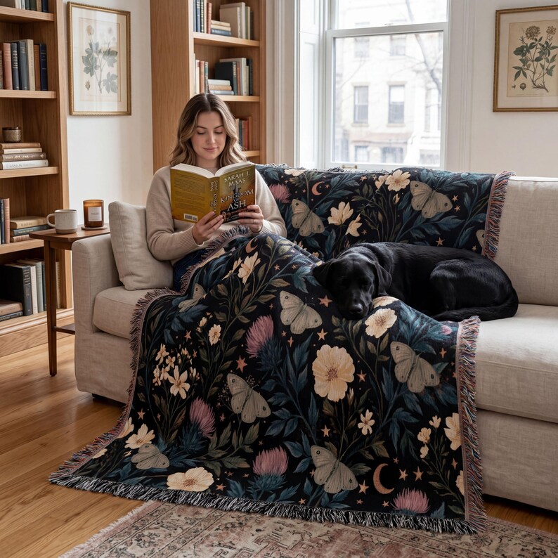 May include: A woven throw blanket featuring a dark background with a repeating pattern of light-colored moths, cream-colored flowers, pink thistle-like blooms, small stars, and crescent moons, finished with a fringed edge on the sides.