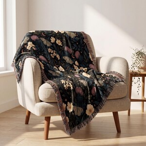 May include: A woven throw blanket featuring a dark background with a repeating pattern of moths, light-colored flowers, pink thistle-like blooms, small stars, and crescent moons. The blanket has a fringed edge and is draped over a light-colored chair.