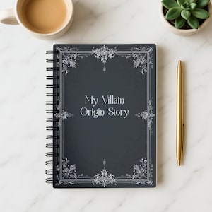 May include: A spiral-bound notebook with the text "My Villain Origin Story" on the cover. The cover is dark gray with ornate silver-colored decorative elements. A gold pen and a cup of coffee are next to the notebook.