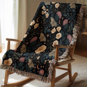 May include: A woven throw blanket featuring a dark background with a pattern of light-colored moths, cream-colored flowers, pink thistle-like blooms, crescent moons, and small stars. The blanket has a fringed edge and is draped over a wooden chair.