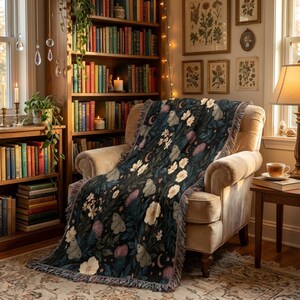 May include: A woven throw blanket featuring a dark background with a botanical pattern of white flowers, pink thistles, and moths, accented by crescent moons and stars, finished with a fringed edge draped over a beige upholstered armchair.