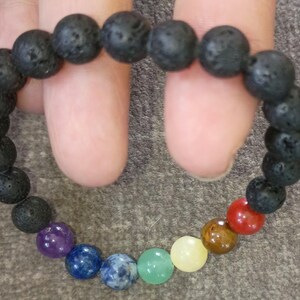 May include: A black lava stone bracelet with a mix of colorful gemstone beads. The bracelet features a series of dark, porous lava beads and a sequence of vibrant, polished gemstones in various colors, including red, orange, yellow, green, blue, and purple.