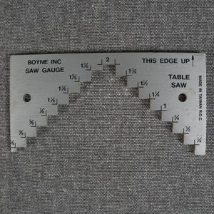 May include: A silver-colored metal Boyne Inc saw gauge for a table saw. The tool features a stepped design with etched markings ranging from 1/8 inch to 2 inches. It includes two circular holes and text reading THIS EDGE UP and MADE IN TAIWAN R.O.C.